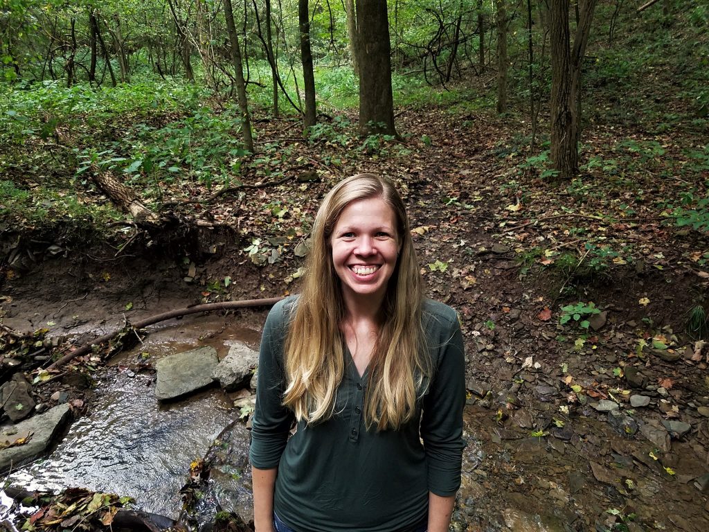 Picture of Maria (blond woman) standing in front of a creek in a wooded area