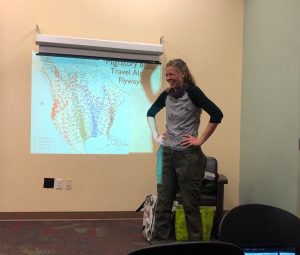 Maria laughing while standing in front of a screen with a PowerPoint presentation that includes a map of migratory flyways in North America